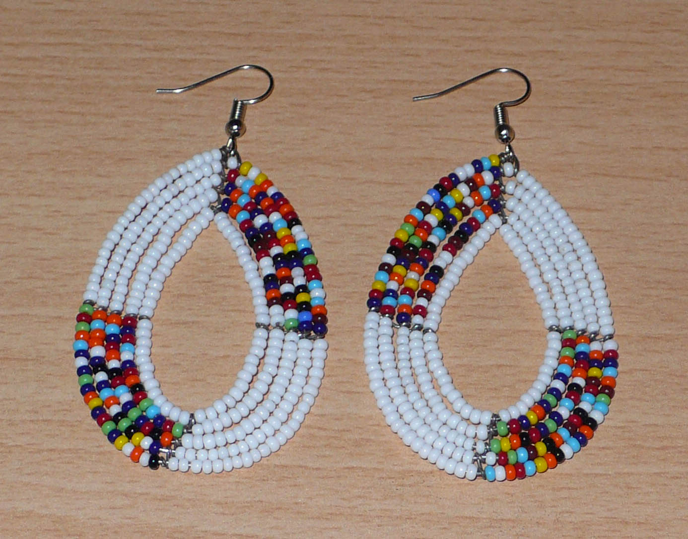 Boucles d'oreilles kenyanes perlées composées de perles de verre blanches et multicolores enfilées sur plusieurs rangées de fils de fer montées en forme de gouttes ovales.  De taille moyenne, ces boucles d'oreilles aux accents exotiques mesurent 7,5 cm de long et 4 cm de large, et se portent avec crochets en acier inoxydable sur des oreilles percées.