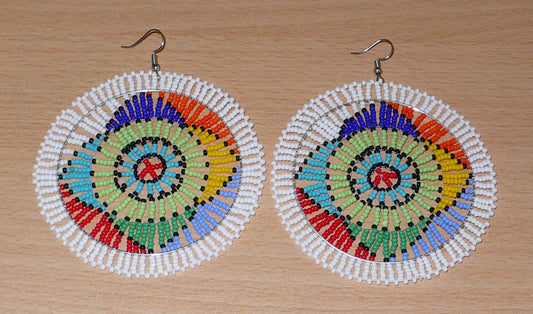 Boucles d'oreilles africaines ethniques, longues et larges, aux couleurs et motifs traditionnels zoulous : elles se composent de perles de rocaille blanches et multicolores enfilées sur des fils de nylon fixés sur des structures circulaires en fils de fer.   Surdimensionnées, ces boucles d'oreilles au style afro chic évoquant les années 70 mesurent 10 cm de long et 8.5 cm de large ; elles se portent avec des crochets en acier inoxydable sur des oreilles percées.