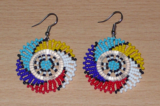 Boucles d'oreilles sud-africaines de forme rondes composées de perles de rocaille aux couleurs exotiques rouges, jaunes, bleu ciel, bleu foncé, blanches et noires glissées sur une trame circulaire faite de plastique et de fils de nylon. Elles mesurent 5,5 cm de long et 4 cm de large. Les crochets d'attache sont en acier inoxydable. Timeless Fineries