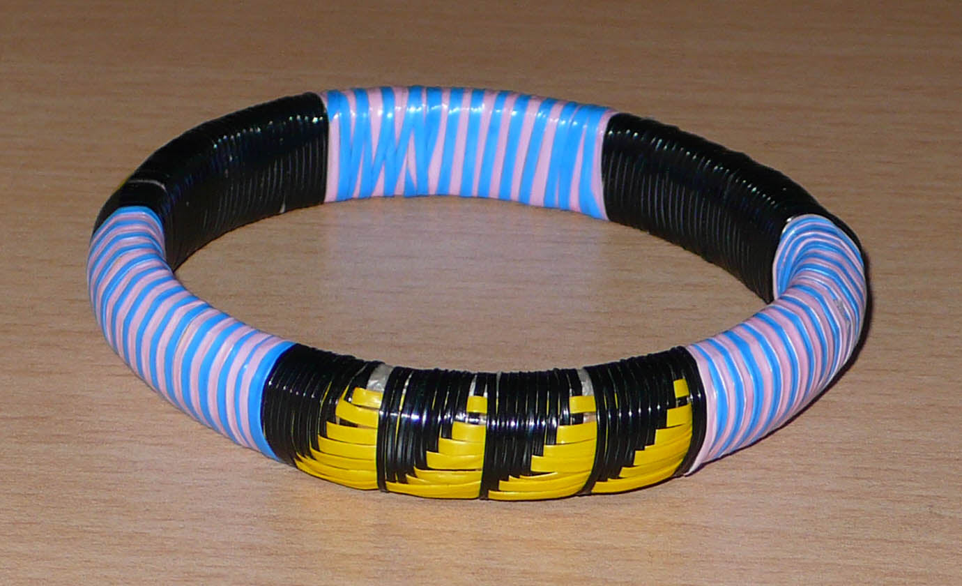 Bracelet africain éco-responsable composé de motifs tissés à partir de fines bandes de plastique recyclé noir, jaune, rose et bleu. La circonférence interne du bracelet est de 20 cm, son diamètre de 6,7 cm et son épaisseur de 1,3 cm. Il peut être porté par une femme ou par un homme.