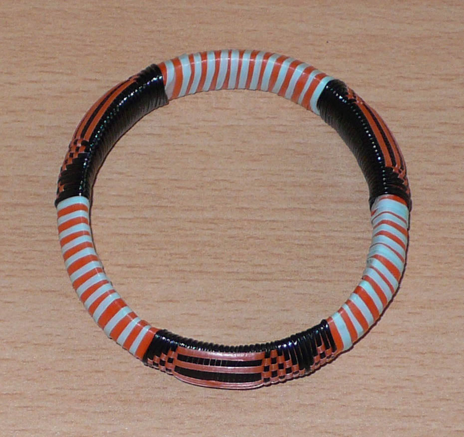 Bracelet africain tribal à motifs ethniques tressés avec du plastique recyclé orange, noir et bleu ciel.

Ce bracelet a une circonférence de 20 cm, un diamètre de 6 cm et une épaisseur de 1.2 cm ; il peut être porté par une femme ou par un homme.