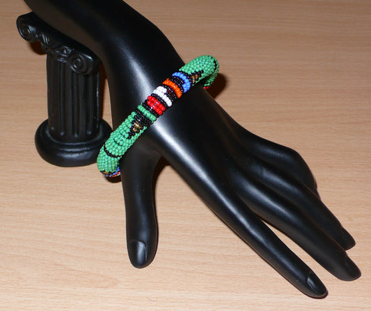 Bracelet africain tubulaire à motifs tribaux zoulous fait de perles de rocaille vertes et multicolores glissées sur du fils de nylon enroulé autour d'un tube en plastique.  Ce bracelet a une circonférence de 21 cm, un diamètre de 6,5 cm et une épaisseur de 1 cm.  Il peut être porté par une femme ou par un homme.