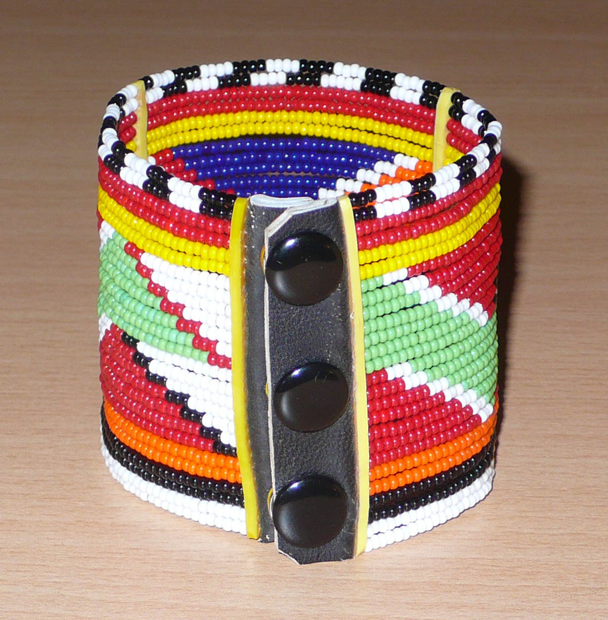 Bracelet africain multi-rangs composé de perles de rocaille aux couleurs et motifs géométriques traditionnels Massai ; les perles multicolores sont enfilées sur vingt-six rangées de fils de fer fixés sur des charnières en plastique. Il s'attache avec des boutons pressions.