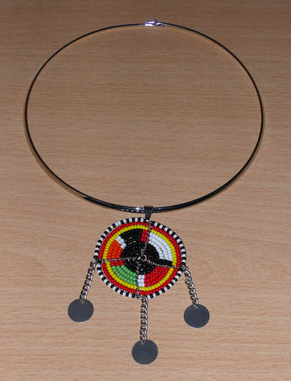 Mélange de tradition Massai et de modernité, ce collier au style tribal chic se compose d'un ras de cou en laiton agrémenté d'un pendentif fait de perles de rocaille noires et multicolores et décoré de petits disques métalliques.  Ce collier mesure 22 cm de long (de la nuque jusqu'à la base du pendentif), la circonférence du ras de cou en laiton est de 42.5 cm avec un diamètre de 13.5 cm ; le pendentif mesure 8 cm de long et 5 cm de large.