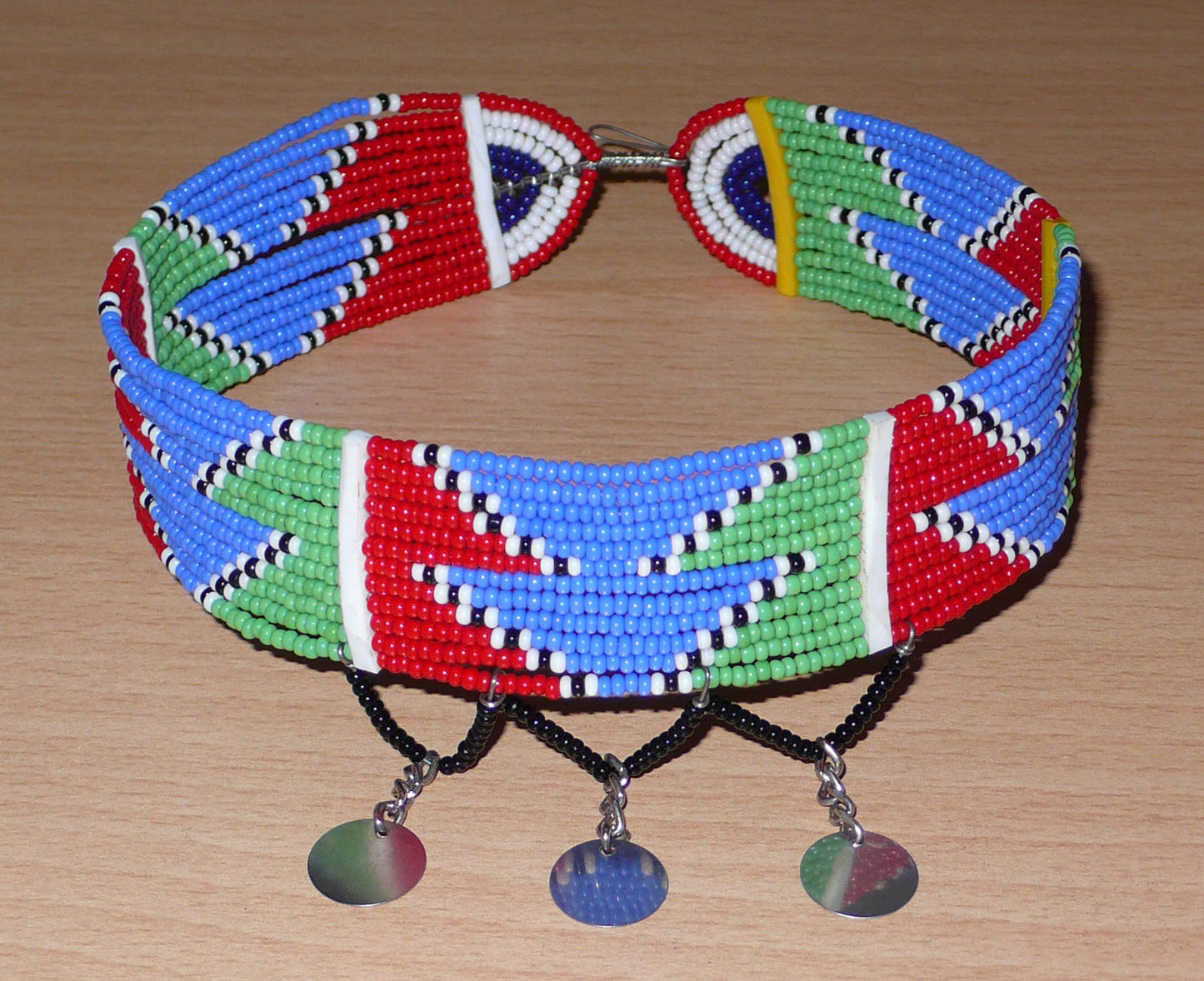 Collier ras de cou africain de tradition Massai composé de douze rangées de perles de rocaille rouges, bleues, vertes, blanches et noires enfilées sur des fils de fer fixés sur des charnières en plastique. Le collier est décoré de petits disques métalliques et se ferme avec un crochet.