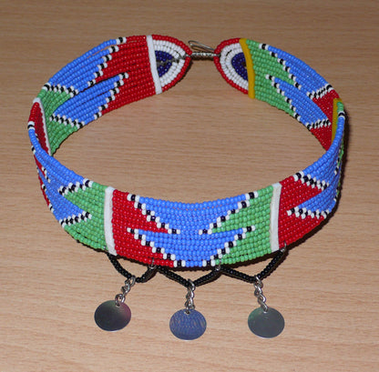 Collier ras de cou africain de tradition Massai composé de douze rangées de perles de rocaille rouges, bleues, vertes, blanches et noires enfilées sur des fils de fer fixés sur des charnières en plastique. Le collier est décoré de petits disques métalliques et se ferme avec un crochet.