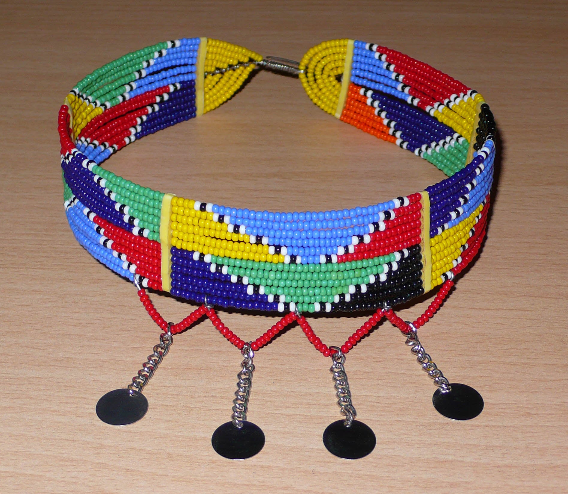 Collier ras de cou africain Massai composé de douze rangées de perles de rocaille multicolores enfilées sur des fils de fer insérés dans des charnières en plastique jaune. Le collier est décoré chaînettes prolongées de petits disques métalliques ; il se ferme avec un crochet.