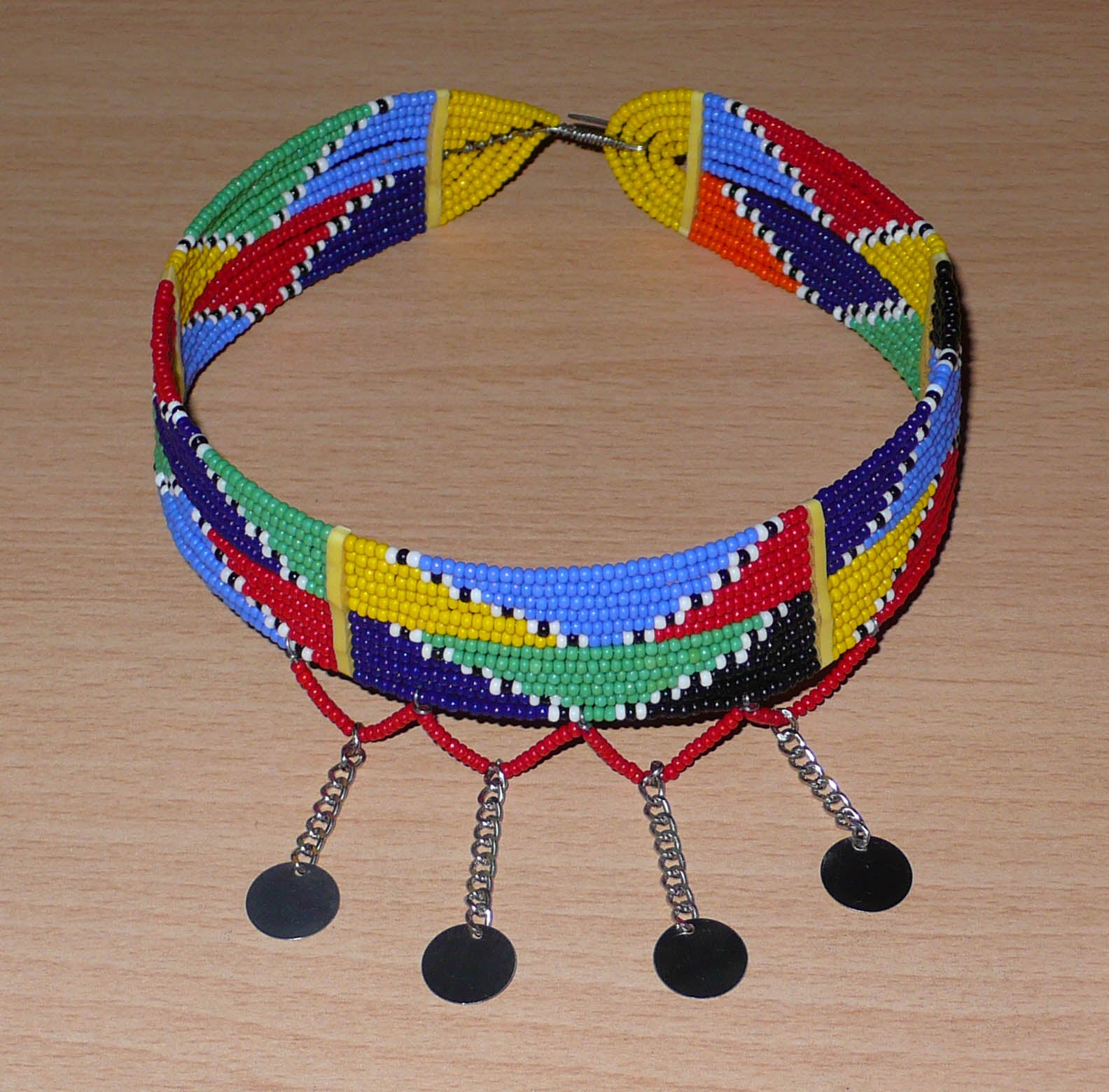 Collier ras de cou africain Massai composé de douze rangées de perles de rocaille multicolores enfilées sur des fils de fer insérés dans des charnières en plastique jaune. Le collier est décoré chaînettes prolongées de petits disques métalliques ; il se ferme avec un crochet.