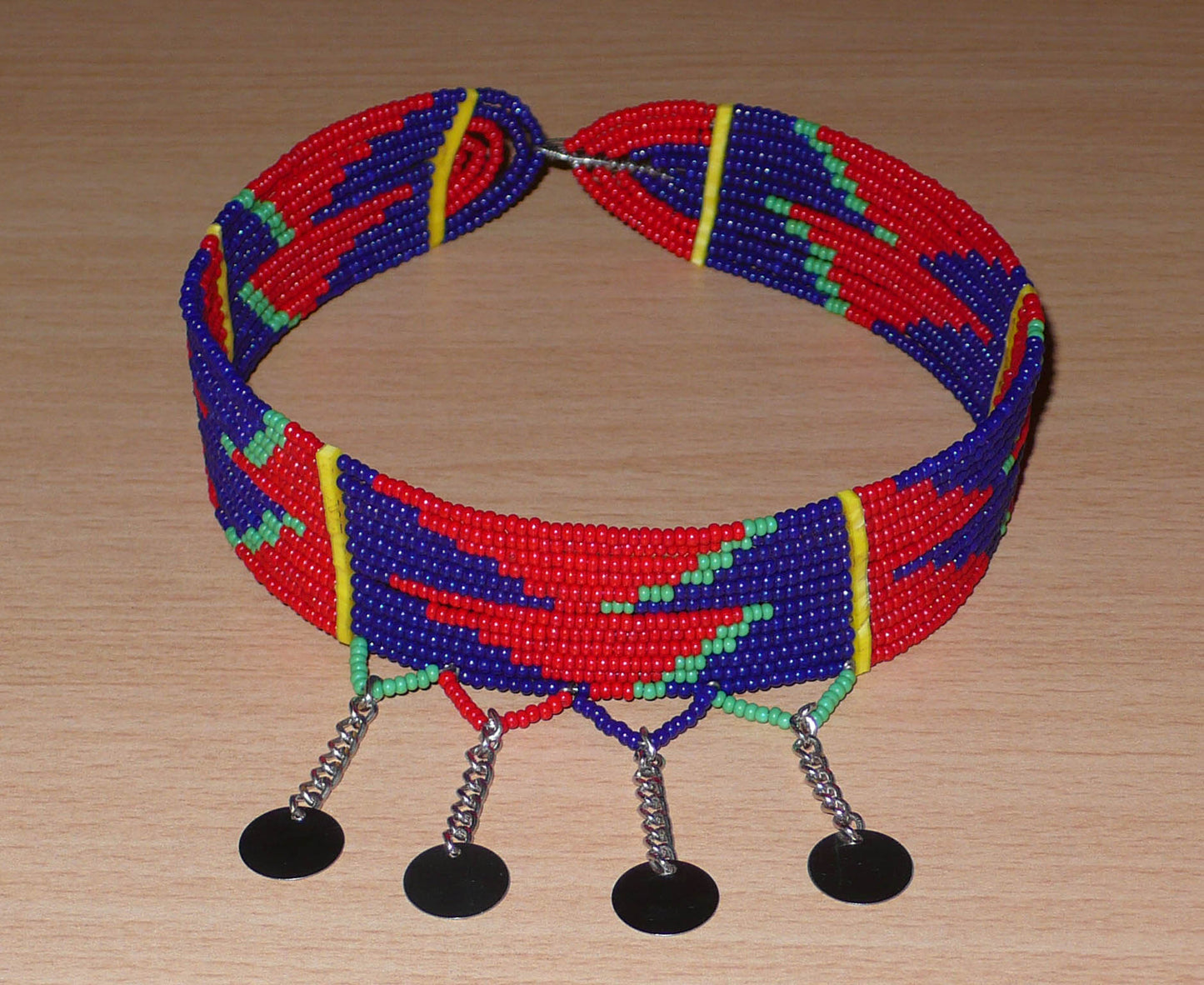 Collier ras de cou africain de tradition Massai composé de douze rangées de perles de rocaille rouges, bleu marine et vertes enfilées sur des fils de fer insérés dans des charnières en plastique jaune. Le collier est décoré chaînettes prolongées de petits disques métalliques ; il se ferme avec un crochet.