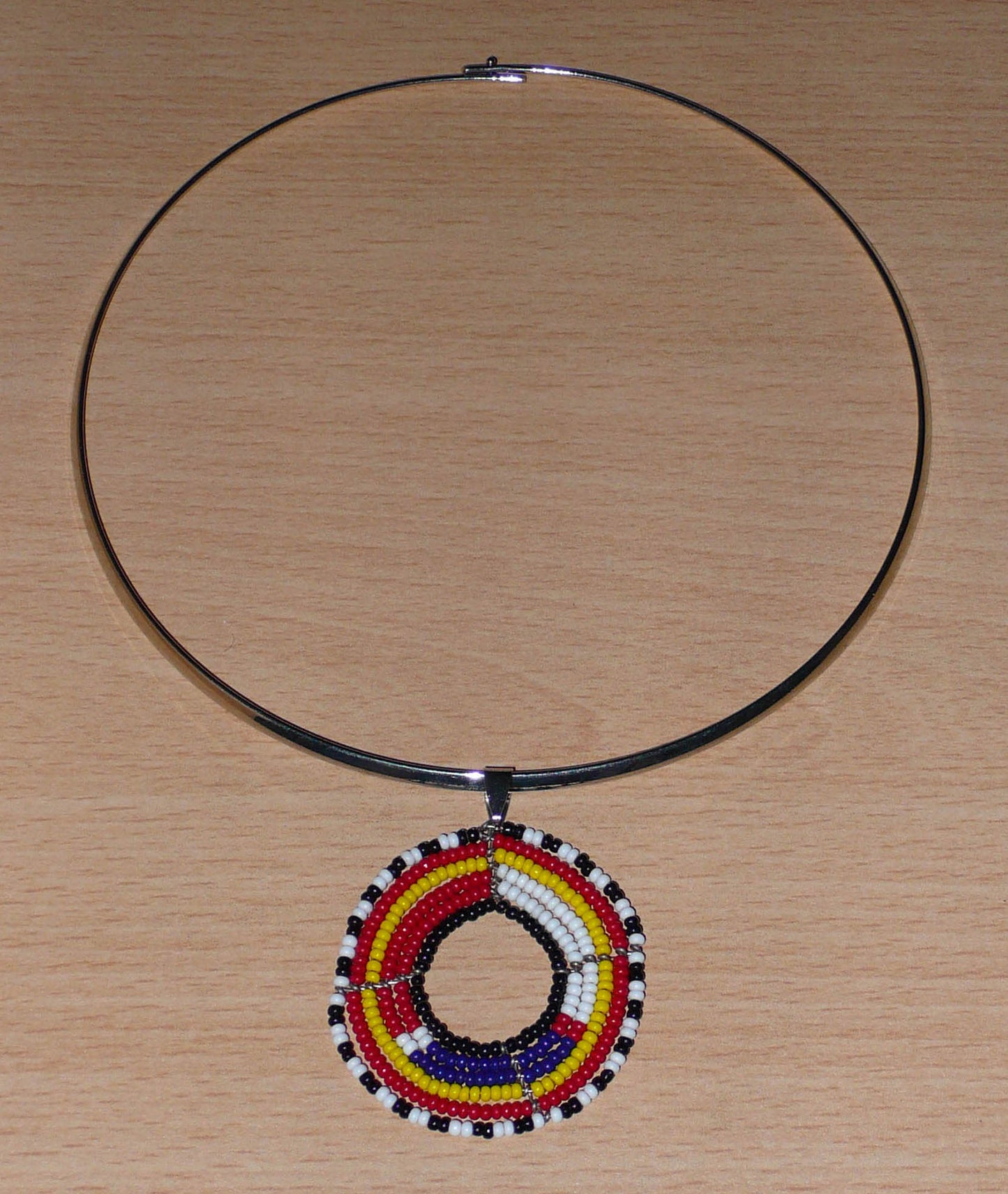 Collier africain contemporain se composant d'un ras de cou en laiton orné d'un pendentif perlé à motifs ethniques Massai fait de perles de rocaille rouges, blanches, bleues, jaunes, noires et blanches glissées sur six rangées de fils de fer montés en cercles.  Ce collier mesure 18.5 cm de long en partant de la nuque jusqu'au pendentif, le ras-de-cou en laiton a une circonférence de 42.5 cm et un diamètre de 13.5 cm ; le médaillon perlé mesure 4.5 cm de long et de large.