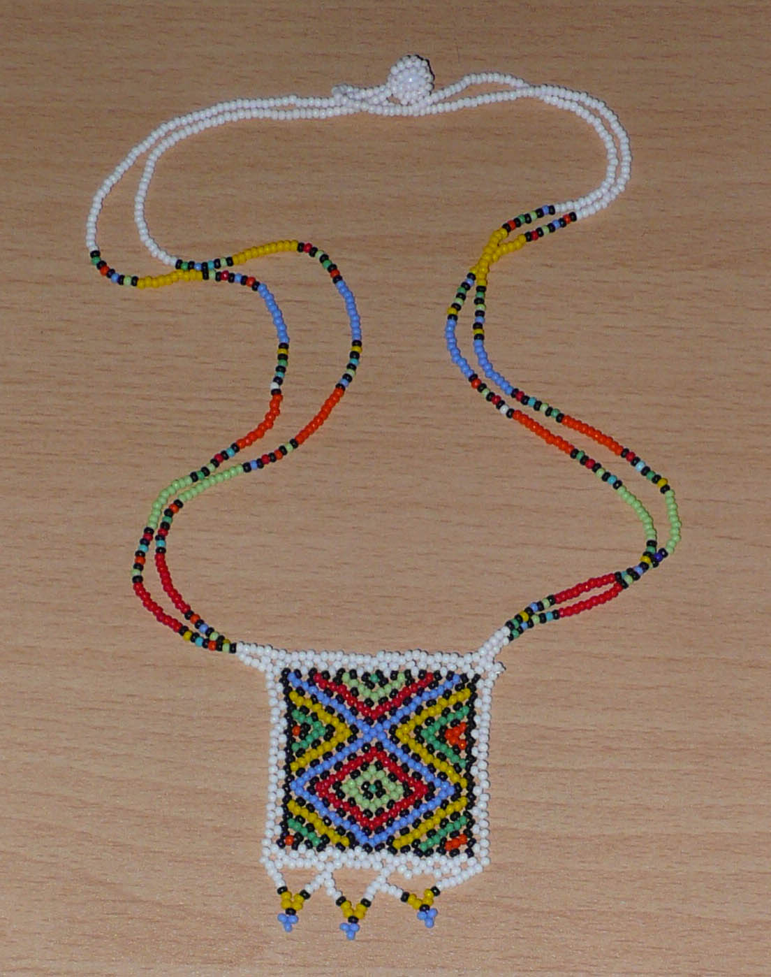 Collier africain traditionnel orné d'un pendentif décoré de motifs ethniques sud-africains ; il se compose d'un tissage de perles de rocaille blanches et multicolores et s'attache avec un fermoir à boule.  Ce collier mesure 28,5 cm de long (de la nuque au pendentif), la longueur de chaîne autour du cou est de 50 cm ; le pendentif carré orné d'une frise perlée mesure 5,5 cm sur 4,5 cm.