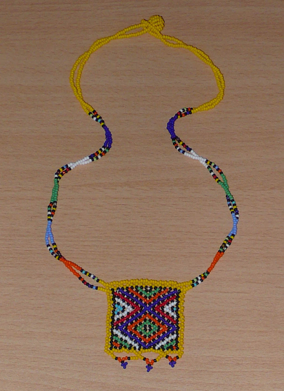 Collier africain orné d'un pendentif carré agrémenté de motifs traditionnels sud-africains. Il se compose de perles de rocaille jaunes et multicolores et s'attache avec un fermoir à boule.  Ce collier mesure 27,5 cm de long (de la nuque au pendentif), la longueur de chaîne autour du cou est de 50 cm ; le pendentif carré orné d'une petite frise perlée mesure 5,5 cm de long et 4,5 cm de large.