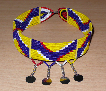 Collier ras de cou ethnique de tradition Massai fait de douze rangées de perles de rocaille jaunes, bleu foncé, rouges et blanches enfilées sur des fils de fer glissés dans des charnières en plastique jaune. Le collier est décoré chaînettes prolongées par des pastilles métalliques ; il se ferme avec un crochet.