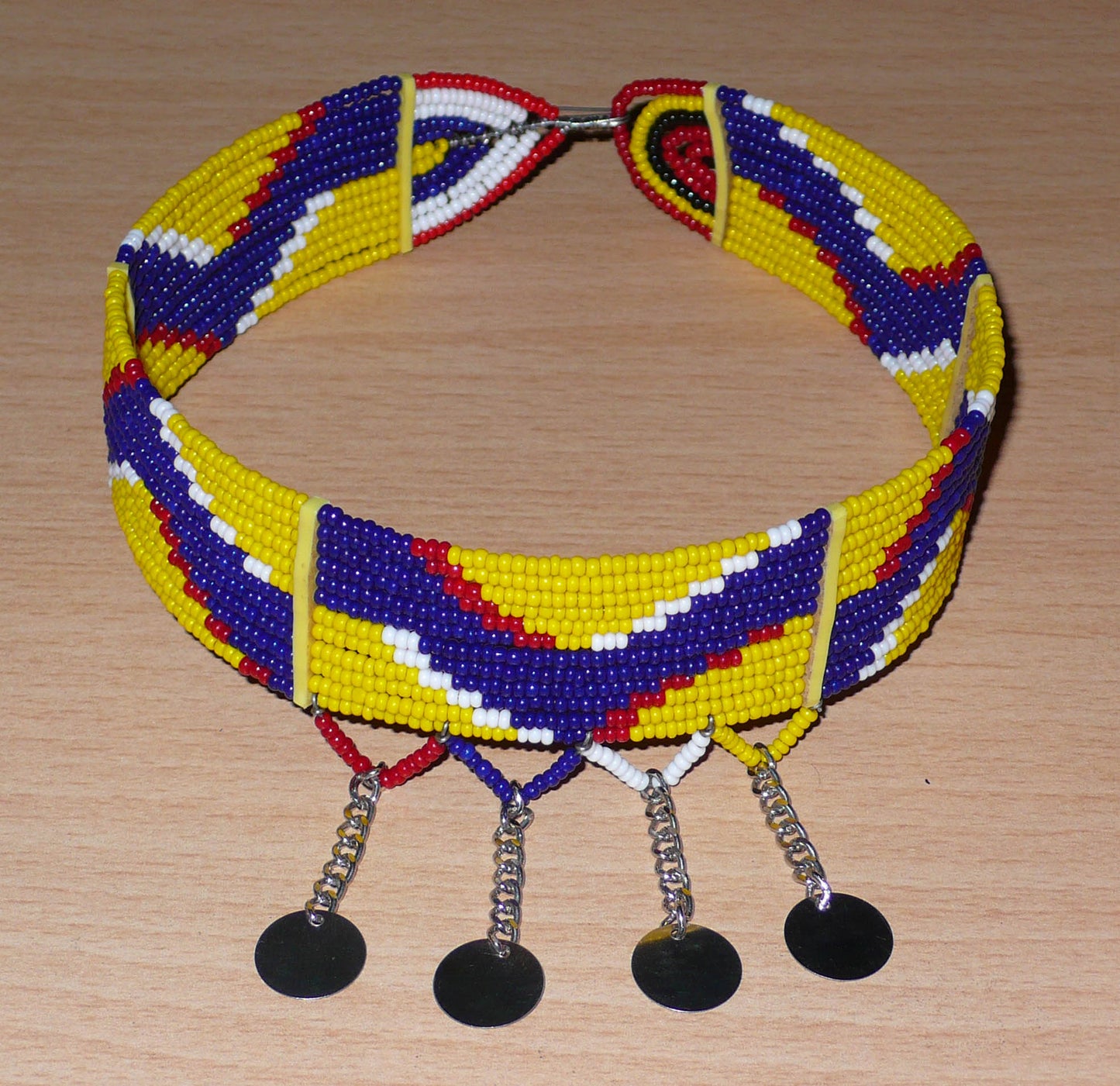 Collier ras de cou ethnique de tradition Massai fait de douze rangées de perles de rocaille jaunes, bleu foncé, rouges et blanches enfilées sur des fils de fer glissés dans des charnières en plastique jaune. Le collier est décoré chaînettes prolongées par des pastilles métalliques ; il se ferme avec un crochet.