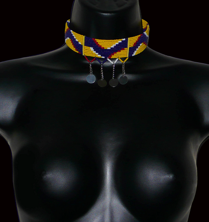 Collier ras de cou ethnique de tradition Massai fait de douze rangées de perles de rocaille jaunes, bleu foncé, rouges et blanches enfilées sur des fils de fer glissés dans des charnières en plastique jaune. Le collier est décoré chaînettes prolongées par des pastilles métalliques ; il se ferme avec un crochet.