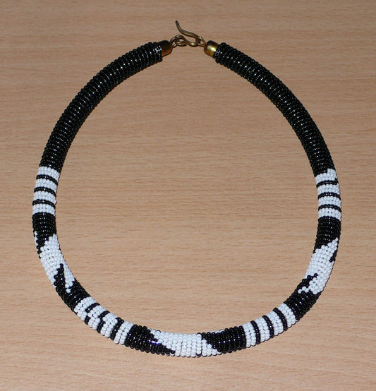 Collier africain tubulaire ras de cou à motifs zoulous composé de perles de rocaille noires et blanches enfilées sur du fil de nylon enroulé autour d'un tube en plastique ; il s'attache avec un crochet. Ce collier ras de cou mesure 17,5 cm de long (de la nuque au bas du collier), la longueur autour du cou est de 46 cm, et son épaisseur est de 1 cm. Ce collier unisexe peut être porté par une femme ou par un homme.