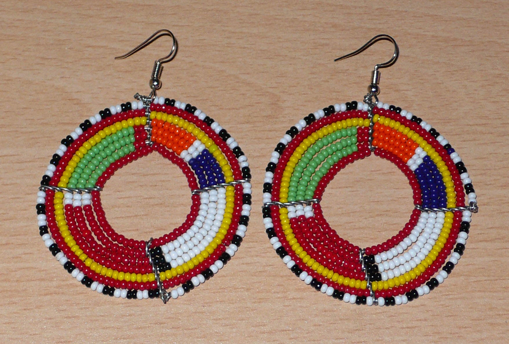 Bijoux africains traditionnels. Boucles d'oreilles africaines rondes aux motifs et couleurs Massai composées de perles de rocaille rouges et multicolores enfilées sur sept rangées de fils de fer montés en forme de cercles. Elles mesurent 7,5 cm de long et 5,5 cm de large et se portent avec des crochets en acier inoxydable sur des oreilles percées. Timeless Fineries