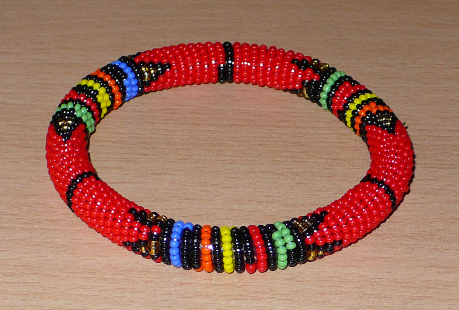 Bracelet africain tubulaire à motifs traditionnels zoulous composé de perles de rocaille rouges et multicolores enfilées sur du fil de nylon enroulé autour d'un tube en plastique.  Ce bracelet a une circonférence interne de 21 cm, un diamètre interne de 6,5 cm et une épaisseur de 1 cm.  Unisexe, il peut être porté par une femme ou par un homme.