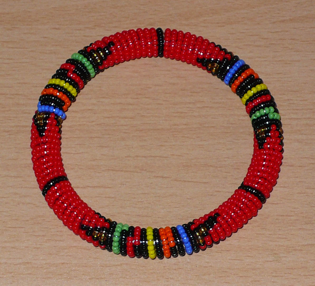 Bracelet africain tubulaire à motifs traditionnels zoulous composé de perles de rocaille rouges et multicolores enfilées sur du fil de nylon enroulé autour d'un tube en plastique.  Ce bracelet a une circonférence interne de 21 cm, un diamètre interne de 6,5 cm et une épaisseur de 1 cm.  Unisexe, il peut être porté par une femme ou par un homme.